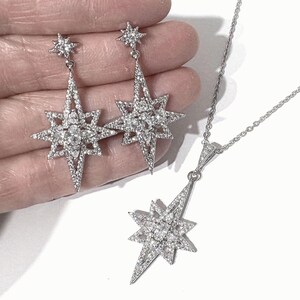 May include: A jewelry set consisting of two drop earrings and a pendant necklace, each featuring a silver-toned starburst design encrusted with clear, sparkling stones. The earrings and pendant share a matching multi-pointed star shape.
