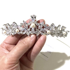 May include: A silver-toned tiara featuring a symmetrical arrangement of clear marquise-cut and round-cut crystals. The headpiece is accented with small, round white pearls interspersed throughout the floral-inspired metal framework.