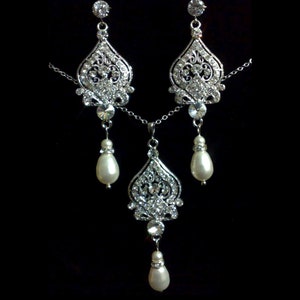 Puede incluir: Un conjunto de collar y pendientes de plata con un colgante de cristal y gotas de perlas. El colgante y los pendientes presentan un dise&ntilde;o delicado y ornamentado.