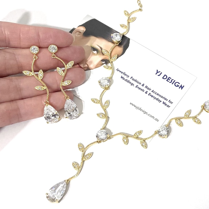 Op de afbeelding: Gouden ketting en oorbellen met heldere kristallen en bladontwerpen. De ketting heeft een lange, hangende hanger met een druppelvormige steen. De oorbellen hebben kleinere druppelvormige stenen.