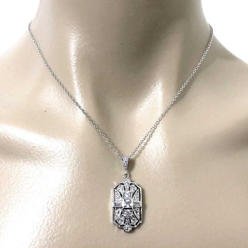 Puede incluir: Un collar de tono plateado con un colgante rectangular de dise&ntilde;o geom&eacute;trico. El colgante est&aacute; engastado con m&uacute;ltiples piedras transparentes facetadas, incluida una piedra central cuadrada rodeada de piedras redondas y de corte baguette.