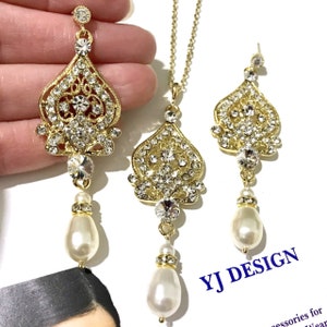 Puede incluir: Conjunto de collar y pendientes en tono dorado con una gota nacarada y un colgante incrustado de cristales. El colgante tiene forma de coraz&oacute;n con intrincados detalles de filigrana. El texto "YJ DESIGN" es visible en la imagen.