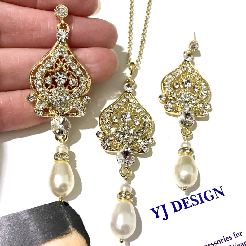 Puede incluir: Conjunto de collar y pendientes en tono dorado con una gota nacarada y un colgante incrustado de cristales. El colgante tiene forma de coraz&oacute;n con intrincados detalles de filigrana. El texto "YJ DESIGN" es visible en la imagen.