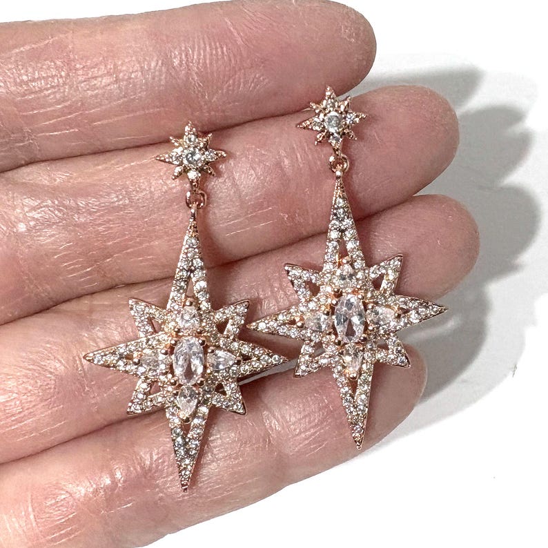 May include: A pair of rose gold-toned drop earrings featuring a starburst design. Each earring consists of a small star-shaped stud connected to a larger, elongated eight-pointed star pendant, both encrusted with clear, sparkling pav&eacute;-set crystals.