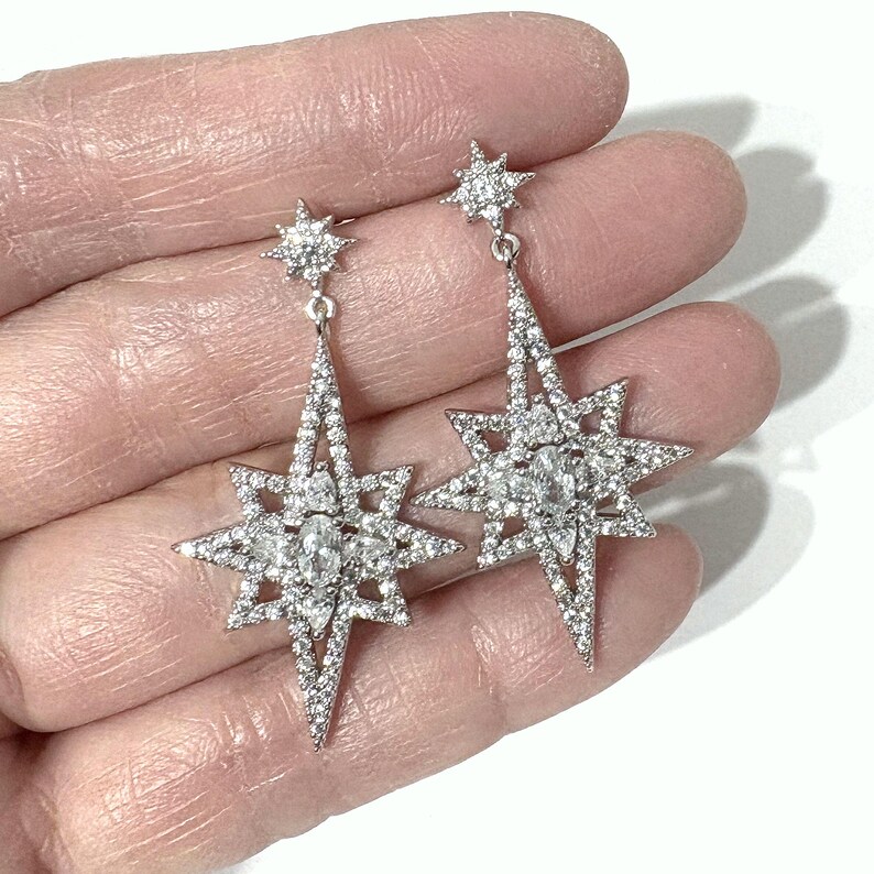 May include: A pair of silver-toned drop earrings featuring a large, multi-pointed star design encrusted with clear, sparkling stones. Each earring is topped with a smaller, matching star stud, creating a cohesive, celestial-inspired jewelry piece.