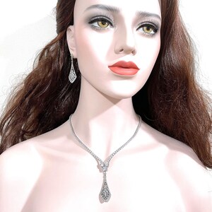 May include: A silver-toned necklace and matching drop earring set featuring clear, faceted stones. The necklace has a V-shaped center with a dangling, elongated, diamond-shaped pendant. The earrings mirror the pendant's shape and intricate design.