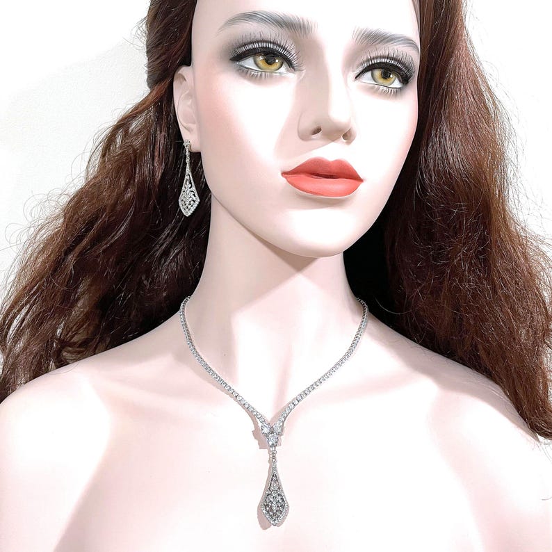 May include: A silver-toned necklace and matching drop earring set featuring clear, faceted stones. The necklace has a V-shaped center with a dangling, elongated, diamond-shaped pendant. The earrings mirror the pendant's shape and intricate design.