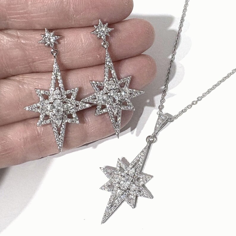 May include: A jewelry set consisting of two drop earrings and a pendant necklace, each featuring a silver-toned starburst design encrusted with clear, sparkling stones. The earrings and pendant share a matching multi-pointed star shape.
