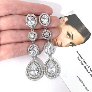 May include: Silver dangle earrings with multiple clear crystal stones in oval and teardrop shapes. The earrings have a layered design with multiple rings of stones.