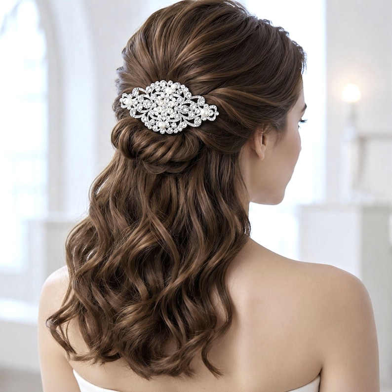 May include: A silver-toned hair clip featuring an intricate, oval-shaped filigree design adorned with numerous clear, sparkling rhinestones and small white pearls. The accessory is secured in long, wavy, brown hair styled in a half-up arrangement.