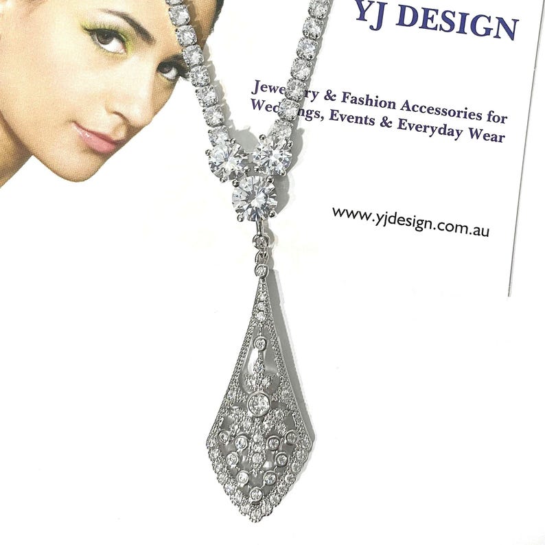 May include: A silver-toned necklace featuring a tennis-style chain with clear round stones leading to a central cluster of two larger stones and a dangling, elongated, diamond-shaped pendant encrusted with small clear stones. Text reads YJ DESIGN.