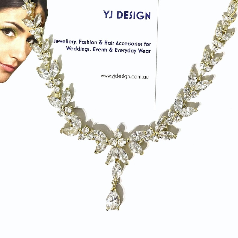 May include: A gold-toned necklace with clear, sparkling gemstones in a leaf and floral design. The necklace has a central pendant and is displayed on a white background. The image also includes text that reads "YJ DESIGN" and "Jewellery, Fashion & Hair Accessories for Weddings, Events & Everyday Wear."