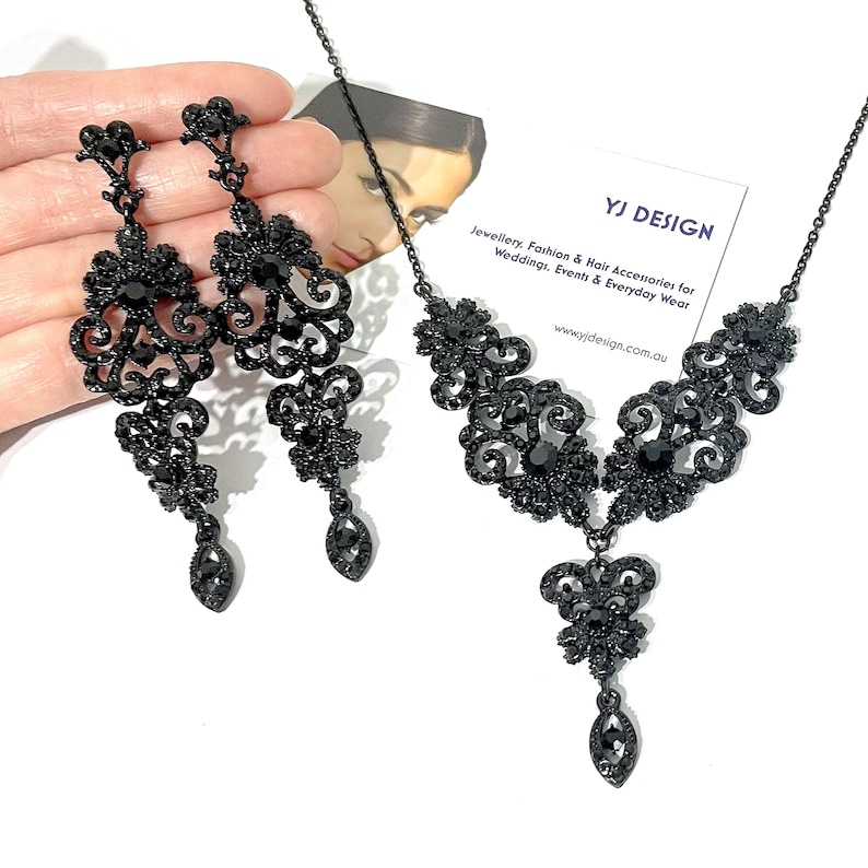 May include: A black jewelry set consisting of a necklace and a pair of long drop earrings. Both pieces feature intricate filigree metalwork adorned with small black crystals and a central teardrop-shaped stone. A YJ DESIGN card is visible.