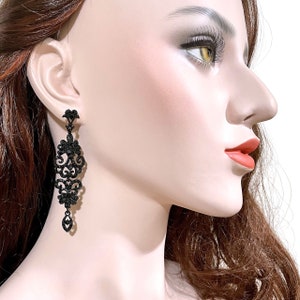 May include: A long, dangling earring featuring an intricate, filigree-style metal design in black. The earring is adorned with numerous small, sparkling black crystals and a single, teardrop-shaped black crystal at the very bottom tip.