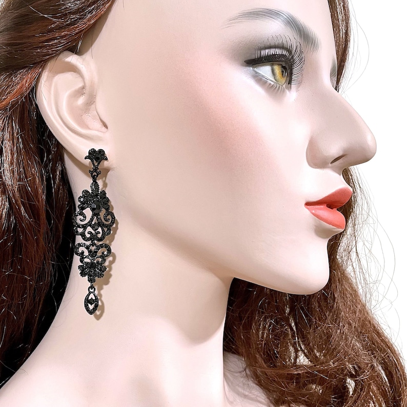 May include: A long, dangling earring featuring an intricate, filigree-style metal design in black. The earring is adorned with numerous small, sparkling black crystals and a single, teardrop-shaped black crystal at the very bottom tip.