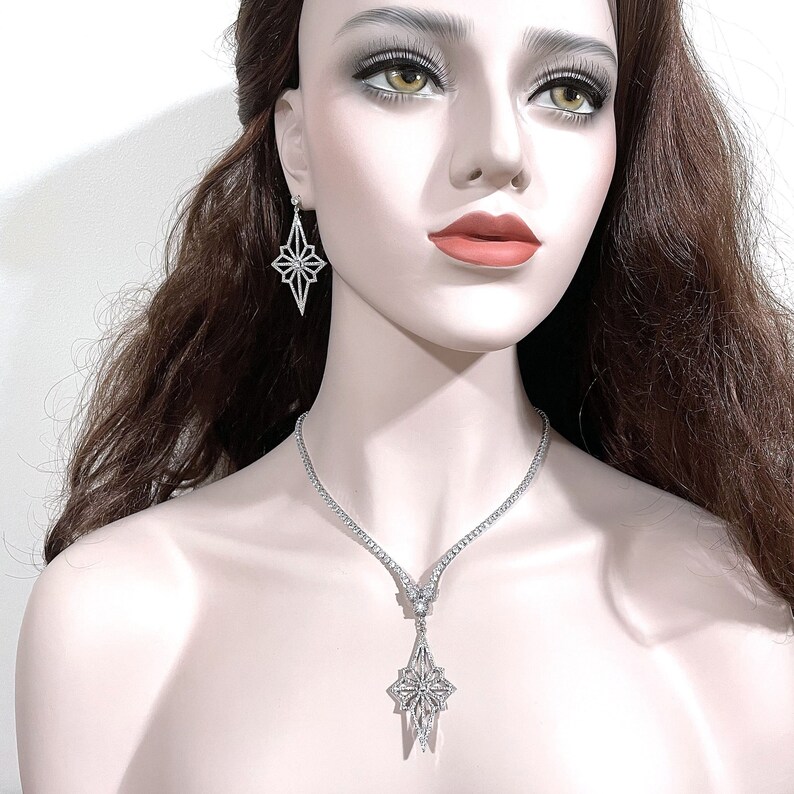 May include: A silver-toned necklace and earring set featuring a starburst or geometric diamond-shaped pendant and matching drop earrings. The necklace consists of a delicate chain with a central star-shaped charm, all encrusted with clear stones.