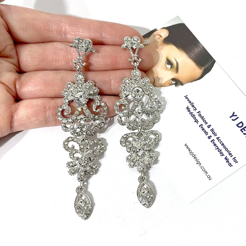 May include: Silver dangle earrings with a floral design and clear rhinestones. The earrings are long and have a teardrop shape at the bottom.