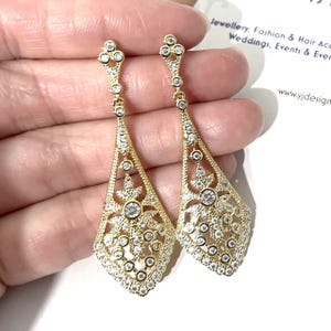 May include: A pair of gold-toned dangle earrings featuring a long, diamond-shaped filigree design. Each earring is encrusted with numerous small, clear round-cut stones and a larger central stone, suspended from a small decorative post.
