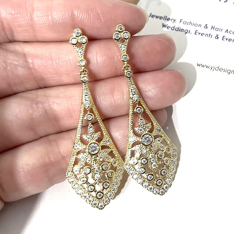 May include: A pair of gold-toned dangle earrings featuring a long, diamond-shaped filigree design. Each earring is encrusted with numerous small, clear round-cut stones and a larger central stone, suspended from a small decorative post.