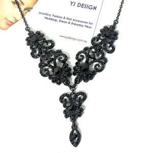 May include: A black necklace featuring intricate filigree metalwork adorned with small black crystals. The design includes symmetrical floral-inspired motifs and a central dangling pendant element, all suspended from a thin black metal chain.