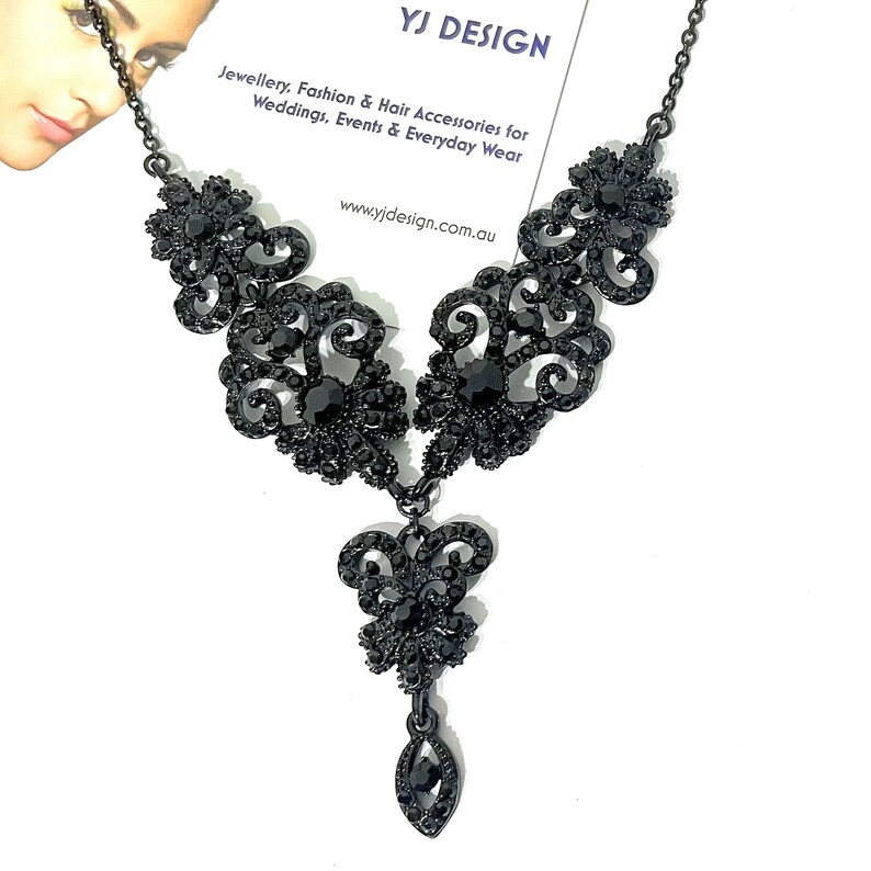 May include: A black necklace featuring intricate filigree metalwork adorned with small black crystals. The design includes symmetrical floral-inspired motifs and a central dangling pendant element, all suspended from a thin black metal chain.