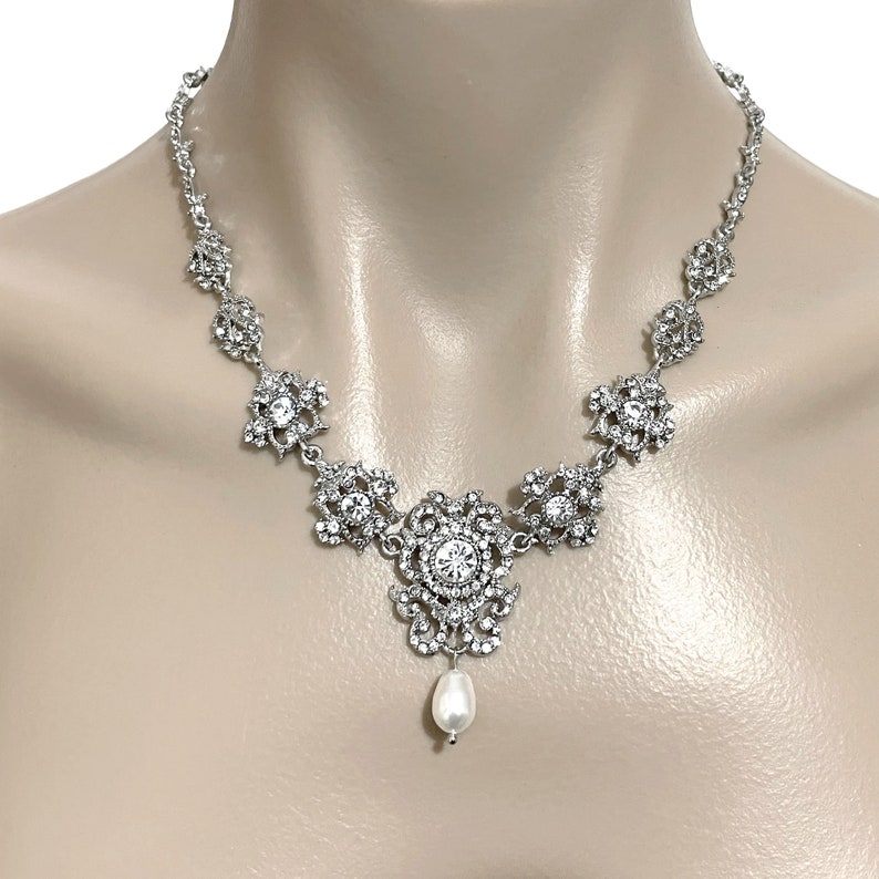 May include: A silver necklace with a large, ornate pendant featuring a pearl drop. The pendant is made of silver and is decorated with clear rhinestones. The necklace is a delicate chain with a similar rhinestone design.