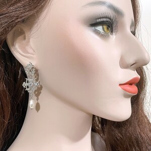 May include: Silver dangle earrings with a pearl drop. The earrings feature a delicate, intricate design with clear rhinestones.