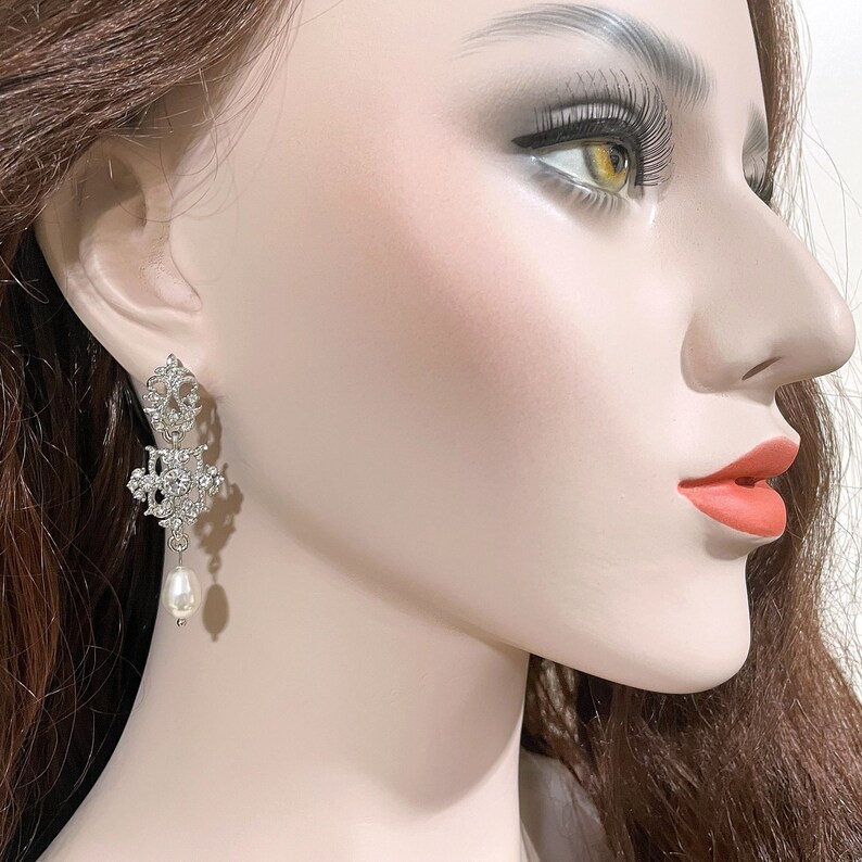 May include: Silver dangle earrings with a pearl drop. The earrings feature a delicate, intricate design with clear rhinestones.