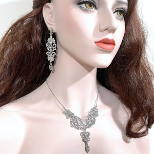 May include: A silver necklace and earrings set with a delicate, intricate design featuring clear rhinestones. The necklace has a pendant that hangs down in the centre.