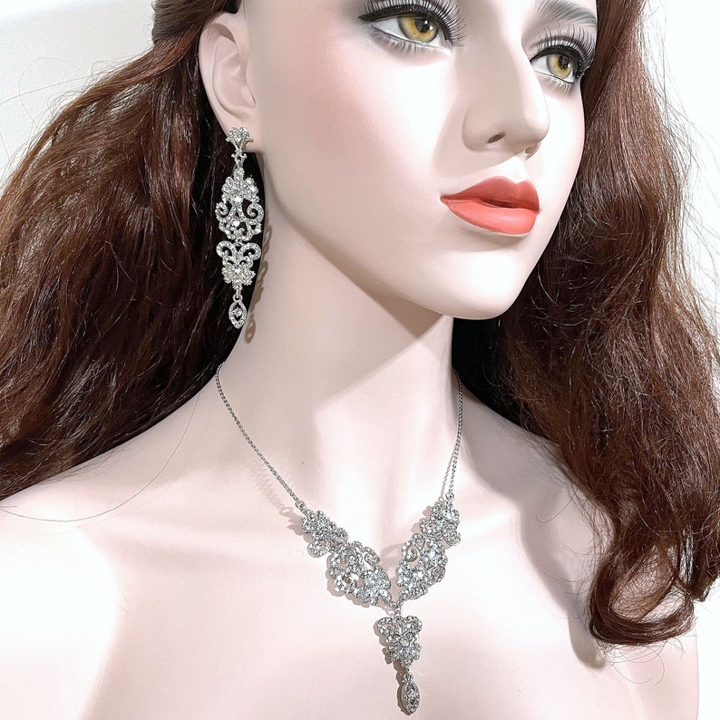 May include: A silver necklace and earrings set with a delicate, intricate design featuring clear rhinestones. The necklace has a pendant that hangs down in the centre.