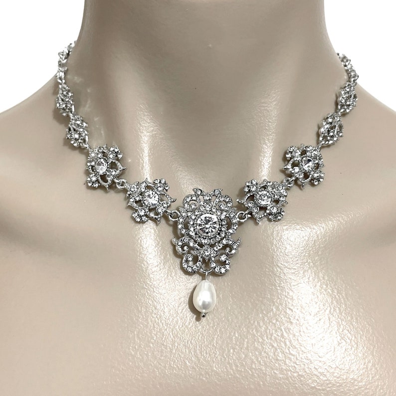 May include: A silver necklace with a large, ornate pendant featuring a pearl drop. The pendant is surrounded by smaller, sparkling stones.