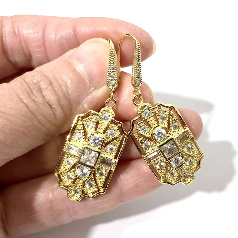 Puede incluir: Un par de pendientes colgantes de tono dorado con un dise&ntilde;o de filigrana rectangular alargado. Cada pendiente est&aacute; incrustado con m&uacute;ltiples piedras de circonita c&uacute;bica transparentes y facetadas en varias formas, incluyendo cortes cuadrados, baguette y redondos.