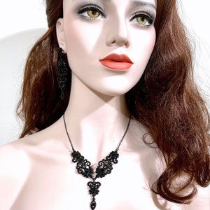 May include: A black necklace and matching earring set featuring intricate filigree metalwork adorned with small black crystals. The necklace has a central drop pendant design, and the earring is a long, dangling piece with a similar ornate pattern.