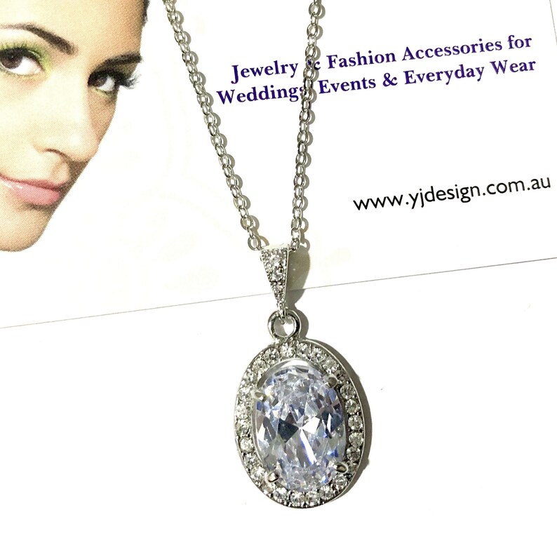 May include: A silver chain necklace with a large oval-shaped pendant featuring a clear cubic zirconia stone surrounded by smaller cubic zirconia stones. The pendant is set in a silver halo setting. The necklace is on a white background with the text "Jewelry & Fashion Accessories for Weddings, Events & Everyday Wear" and "www.yijdesign.com.au".