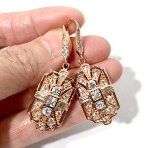 Puede incluir: Un par de pendientes colgantes de oro rosa con un dise&ntilde;o geom&eacute;trico intrincado y piedras transparentes facetadas. Los pendientes incluyen una piedra cuadrada central, detalles en corte baguette y peque&ntilde;as piedras redondas.