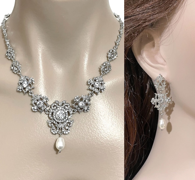 May include: A silver necklace with a large, ornate pendant featuring a pearl drop. Matching earrings with a pearl drop dangle from silver filigree.