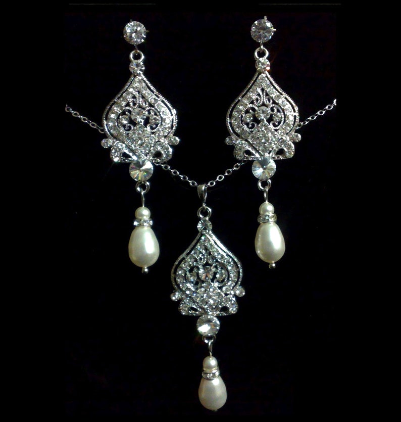 Puede incluir: Un conjunto de collar y pendientes de plata con un colgante de cristal y gotas de perlas. El colgante y los pendientes presentan un dise&ntilde;o delicado y ornamentado.