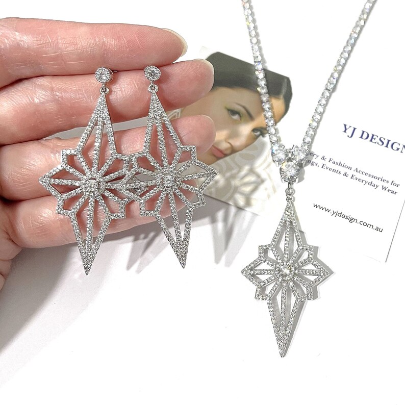May include: A silver-toned jewelry set consisting of two dangling earrings and a matching necklace. Each piece features a geometric starburst design encrusted with clear stones. The necklace includes a tennis-style chain with a central pendant.