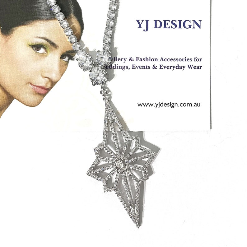 May include: A silver-toned necklace featuring a row of clear round stones leading to a large, geometric star-shaped pendant encrusted with small clear stones. The necklace is displayed next to a card with the text YJ DESIGN, Jewellery & Fashion Accessories for weddings, Events & Everyday Wear, and www.yjdesign.com.au.