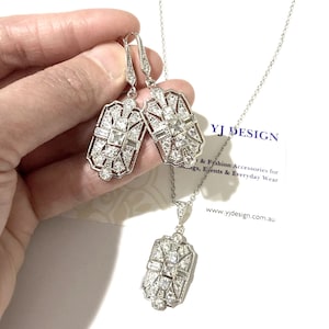 Puede incluir: Un conjunto de joyer&iacute;a en tono plateado que consta de dos pendientes rectangulares y un collar con colgante a juego. Cada pieza presenta un marco de metal geom&eacute;trico incrustado con piedras claras facetadas. Texto YJ DESIGN visible.