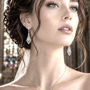 May include: A pair of silver-toned drop earrings featuring a delicate, elongated diamond-shaped filigree design with small clear stones. The earrings hang from a simple post setting, showcasing an intricate openwork pattern that tapers to a point.