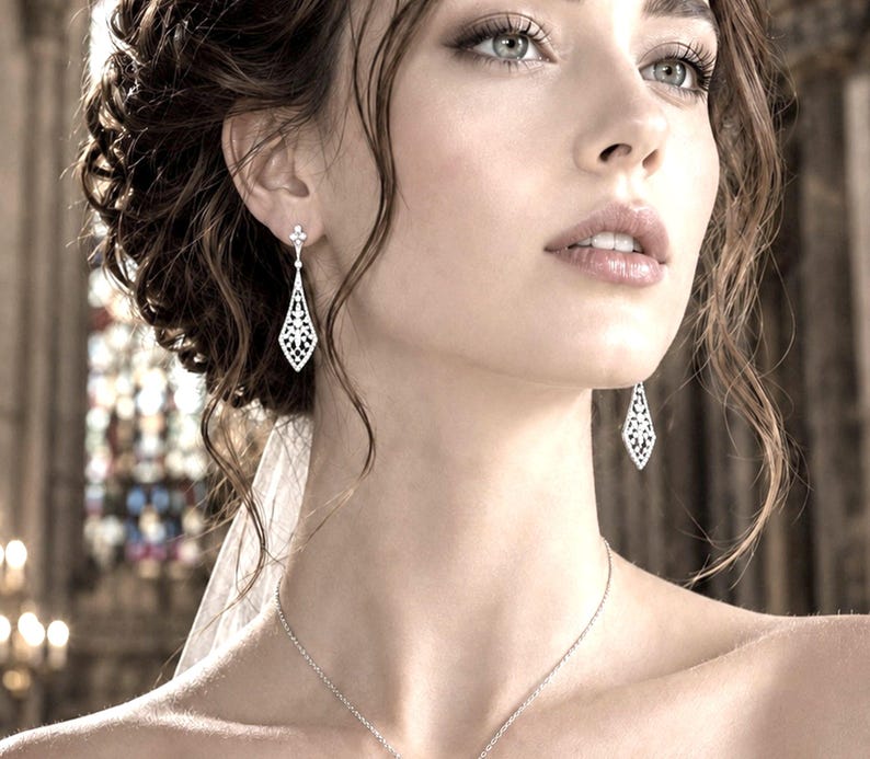 May include: A pair of silver-toned drop earrings featuring a delicate, elongated diamond-shaped filigree design with small clear stones. The earrings hang from a simple post setting, showcasing an intricate openwork pattern that tapers to a point.