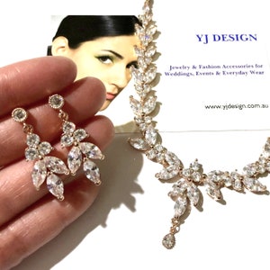 May include: A rose gold jewelry set featuring a necklace and earrings. The necklace and earrings have a leaf design with clear, sparkling crystals. The earrings are dangling. The jewelry is displayed with a business card that reads "YJ DESIGN" and "Jewelry & Fashion Accessories for Weddings, Events & Everyday Wear."