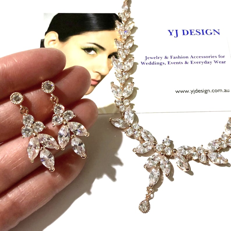 May include: A rose gold jewelry set featuring a necklace and earrings. The necklace and earrings have a leaf design with clear, sparkling crystals. The earrings are dangling. The jewelry is displayed with a business card that reads "YJ DESIGN" and "Jewelry & Fashion Accessories for Weddings, Events & Everyday Wear."