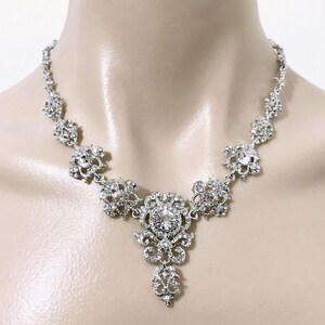 May include: A silver necklace with a large, ornate pendant featuring a clear crystal in the center. The pendant is surrounded by smaller crystals and has a delicate, filigree design.