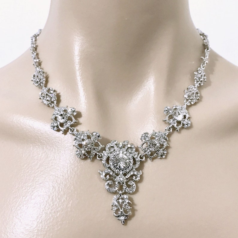 May include: A silver necklace with a large, ornate pendant featuring a clear crystal in the center. The pendant is surrounded by smaller crystals and has a delicate, filigree design.