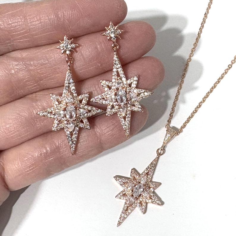 May include: A rose gold jewelry set consisting of two drop earrings and a pendant necklace. Each piece features a multi-pointed star design encrusted with small clear stones and a central oval stone, suspended from a delicate rose gold chain.