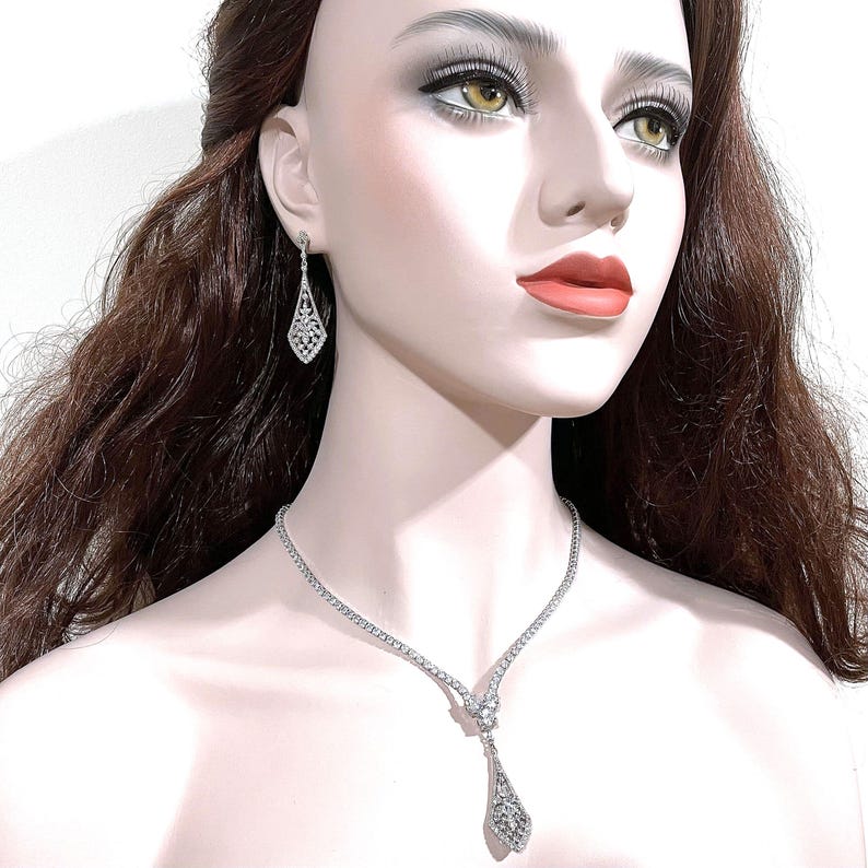 May include: A silver-toned necklace and matching drop earrings set featuring clear, faceted stones. The necklace has a delicate chain with a central pendant, while the earrings have a matching elongated, diamond-shaped design with intricate stone detailing.