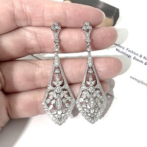 May include: A pair of silver-toned drop earrings featuring an elongated, diamond-shaped filigree design. The metalwork is encrusted with numerous small, clear, round-cut stones, creating a sparkling, intricate pattern that tapers to a point.