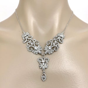 May include: A silver necklace with a delicate, ornate design featuring a large, teardrop-shaped pendant. The pendant is adorned with sparkling clear rhinestones.